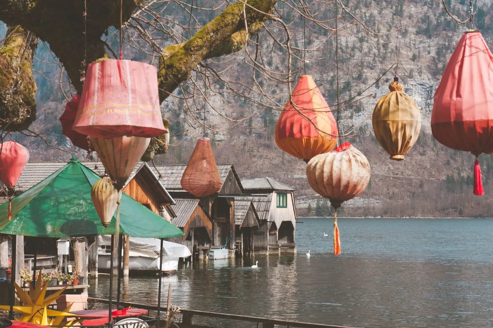 Colorful lanterns hanging over a serene lake in Hallstatt, Austria, with rustic boathouses in the background.